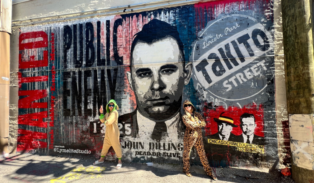 two people in halloween costumes in front of a mrual in dillinger alley on a chicago crime tour